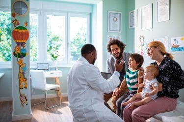 Healthcare professional sits with two adults and two children in medical office exam room. Doctor is holding a tablet-like device in one hand and looks to be explaining something while the group listens.